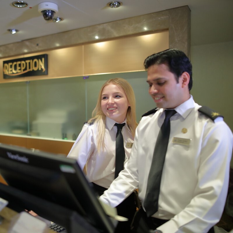 Two hotel receptionists in uniform stand behind a desk, smiling and looking at a computer screen. A Reception sign is visible in the background, indicating they are in the hotel foyer.