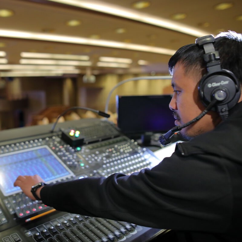 A person wearing a headset operates a professional audio mixing desk in a large, well-lit room, focusing on adjusting the controls.