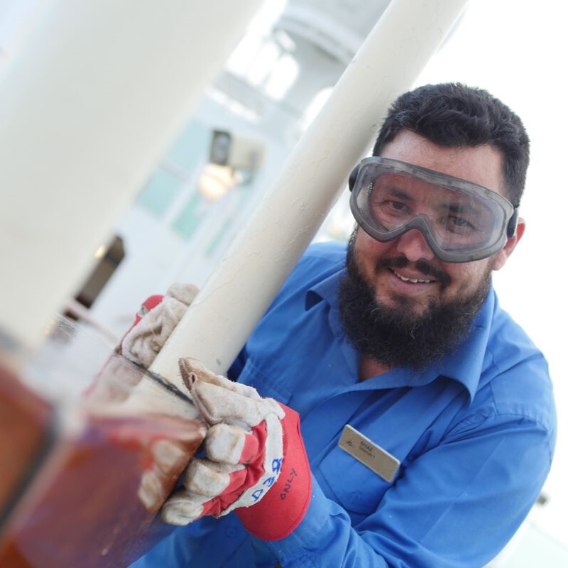 A man wearing safety goggles, gloves, and a blue shirt with a name badge smiles whilst working outdoors as part of the P&O Cruises deck careers team, holding onto a white metal pole.