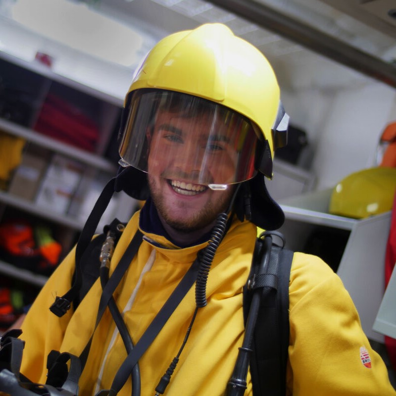 A smiling person in a yellow helmet, face shield, and protective suit stands indoors—perhaps training for P&O Cruises deck careers. Equipment and shelves are visible in the background, suggesting an emergency services environment.
