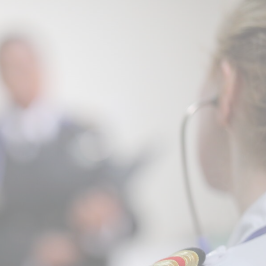 A close-up view of a person in uniform, possibly military, with a blurred background showing indistinct figures. The image is softly focused, highlighting the uniform's shoulder details.