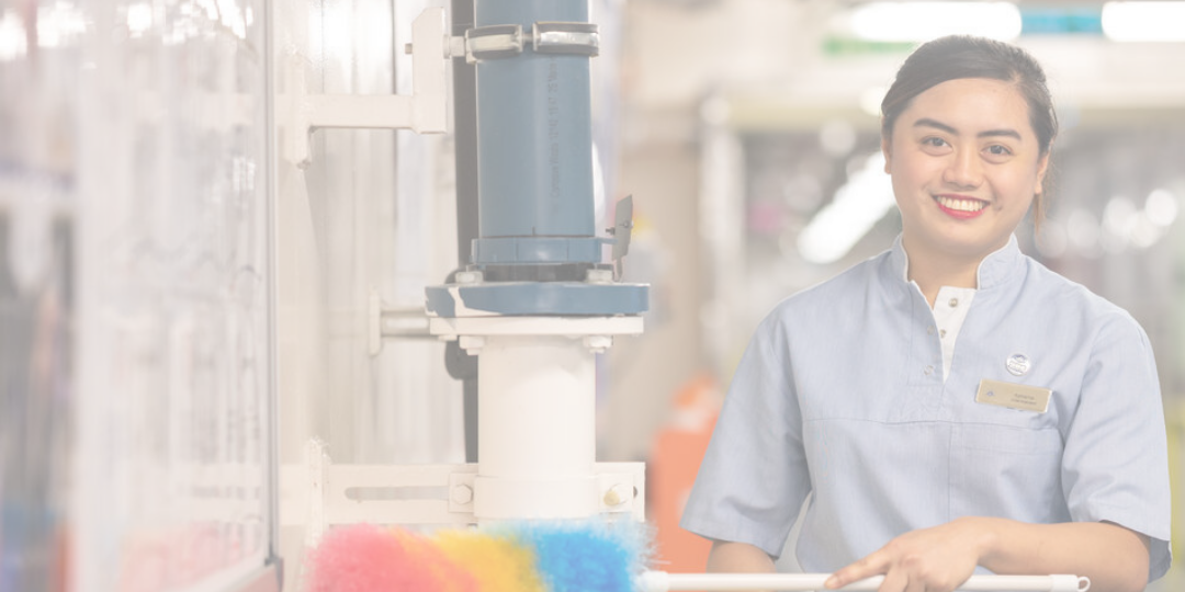 A woman in a light blue uniform is smiling whilst holding a colourful duster in a clean, indoor environment.