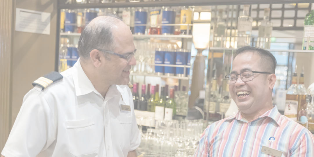 Two men, one in a white uniform and the other in a striped shirt, stand in front of a bar with bottles and glasses, smiling and talking to each other.