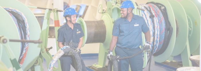 Two crew members in blue uniforms and helmets smile whilst handling ropes on a ship's deck, surrounded by large green winches and coiled ropes—a snapshot of daily life in P&O Cruises deck careers.