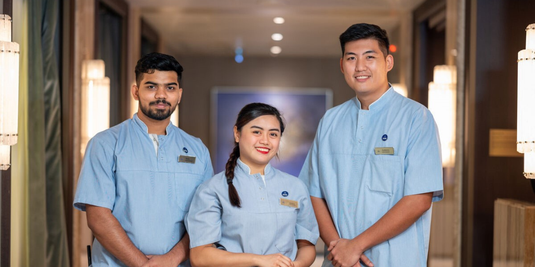 Three hospitality workers wearing light blue uniforms and name badges stand smiling in a well-lit corridor with warm lighting and modern decor.