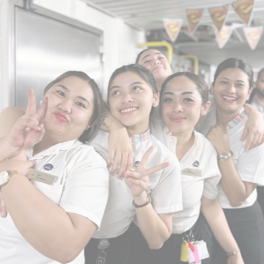 Five smiling women in uniform pose together, some making peace signs with their hands. They are standing indoors, close to each other, appearing cheerful and friendly.