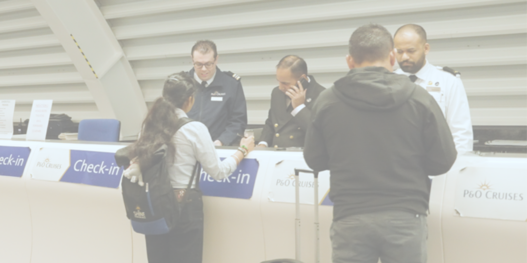 People are checking in at a P&O Cruises counter. Two staff in uniform are assisting guests; one is on the phone. Two people with luggage stand at the counter. Signs reading Check-in are visible.