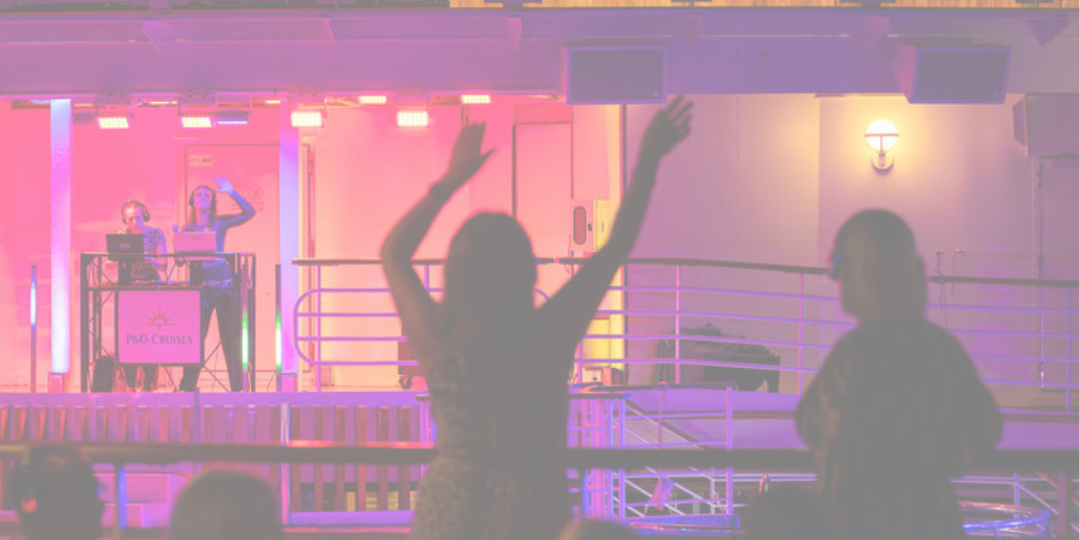 People dance and raise their arms in front of a DJ booth on a cruise ship deck, with colourful lights illuminating the scene.