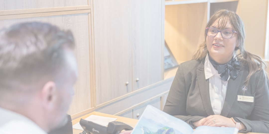 A woman in professional attire sits at a desk, smiling and speaking with a man who is looking at a brochure. The setting appears to be an office or reception area with wooden cupboards in the background.