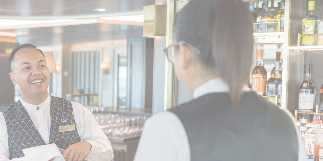 Two bartenders in uniform smile and chat behind a well-stocked bar, surrounded by shelves of spirits bottles. Perfect for those interested in cruise ship bar jobs, one bartender holds a tea towel while the other faces away from the camera.
