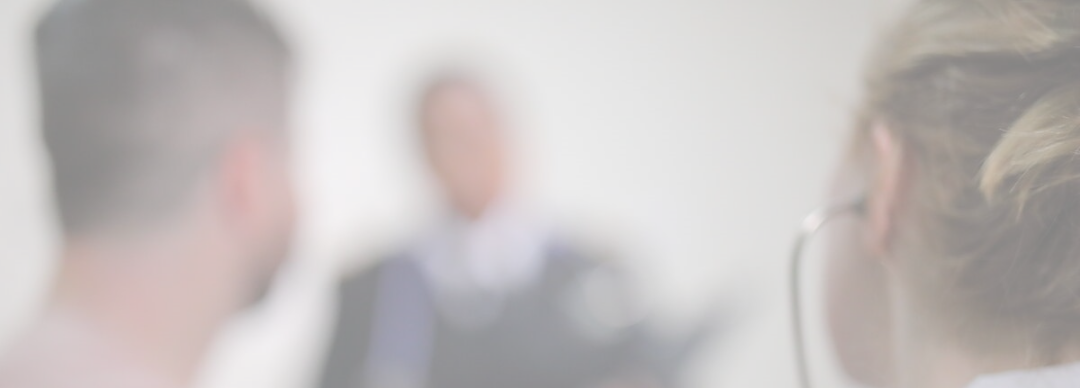 Three people appear blurry in the image; two are in the foreground facing away from the camera, whilst one person in formal attire is in the background, possibly speaking or presenting. The scene is softly lit and out of focus.