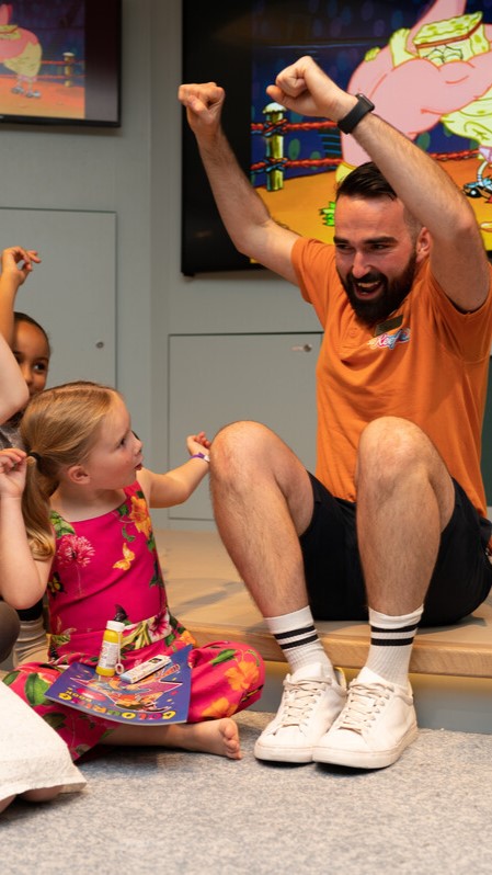 A bearded man in an orange shirt sits on the floor with children, smiling and raising his arms in excitement. A young girl in a pink floral dress sits beside him, holding a glue stick and looking up at him, while cartoons play on a screen behind them.