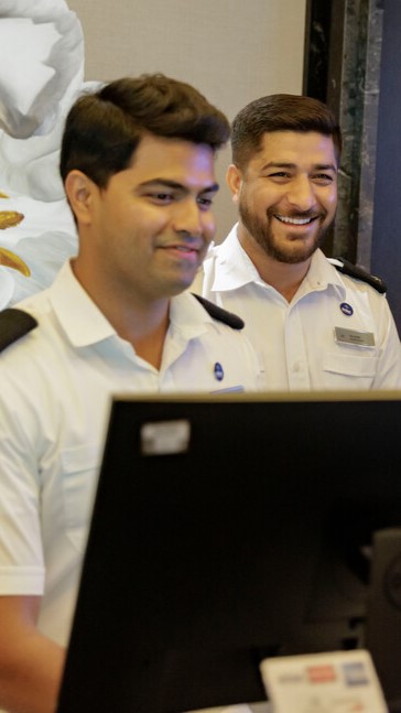 Two smiling men in white uniforms stand behind a desk with a computer monitor, suggesting they are hotel or cruise staff. A decorative floral wall art is partially visible in the background.