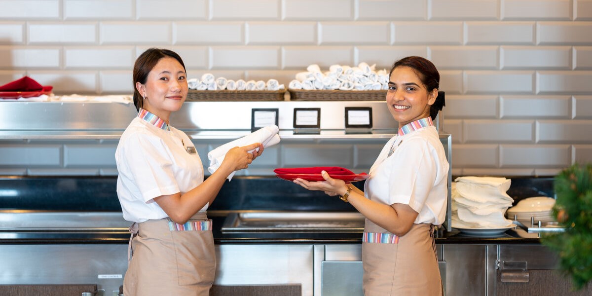 Two women wearing uniforms and aprons stand in a kitchen, smiling at the camera—one holds a rolled white towel, the other a stack of red serviettes. Clean towels are stacked behind them, showcasing team spirit found in cruise ship restaurant jobs.