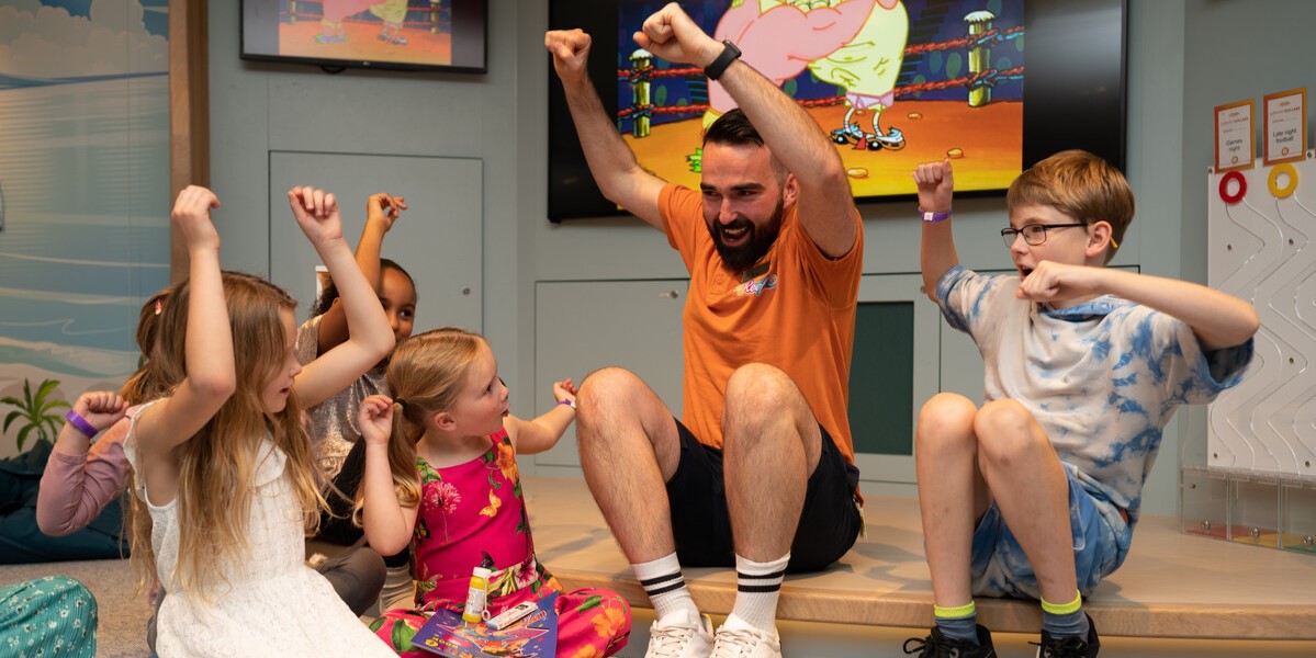 A man sits on the floor with a group of smiling children, all raising their arms excitedly. Colourful cartoon images are displayed on screens behind them, and the group appears to be having fun together in a lively, indoor setting.