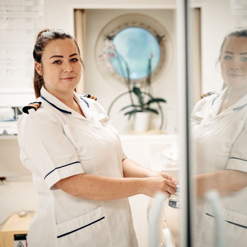 A nurse in a white uniform stands smiling beside a counter, with a round window and a potted orchid in the background. Her reflection appears in a glass surface nearby.