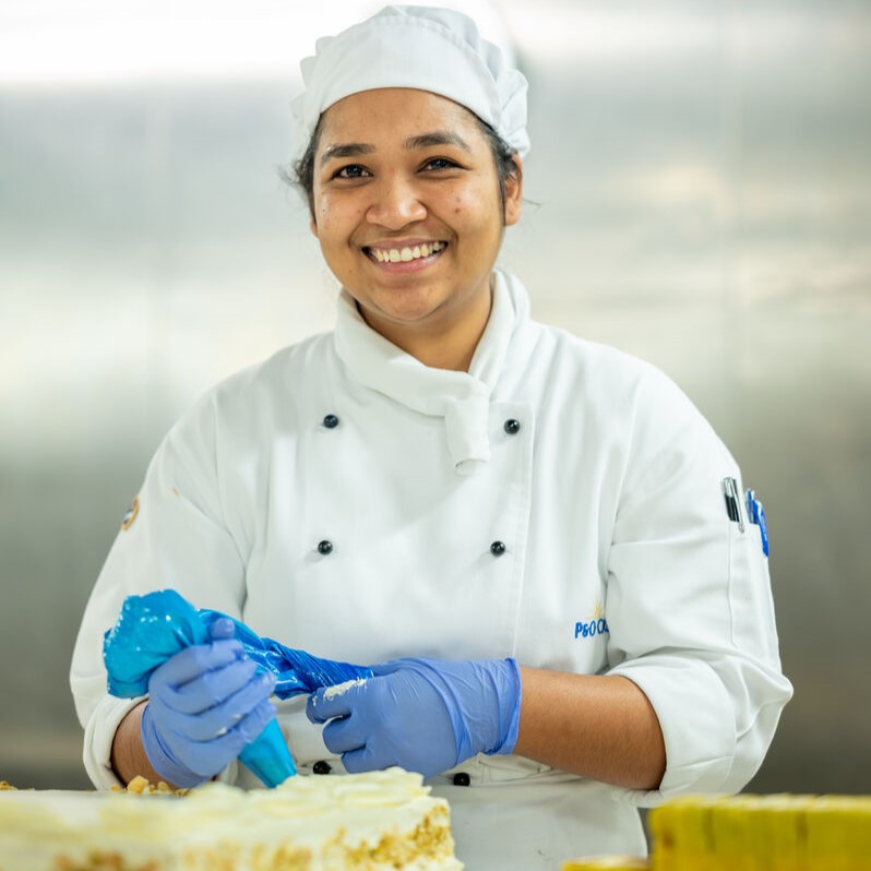 A smiling pastry chef in a white uniform and hat decorates a cake with blue piping bags while wearing blue gloves in a professional cruise ship galley kitchen.