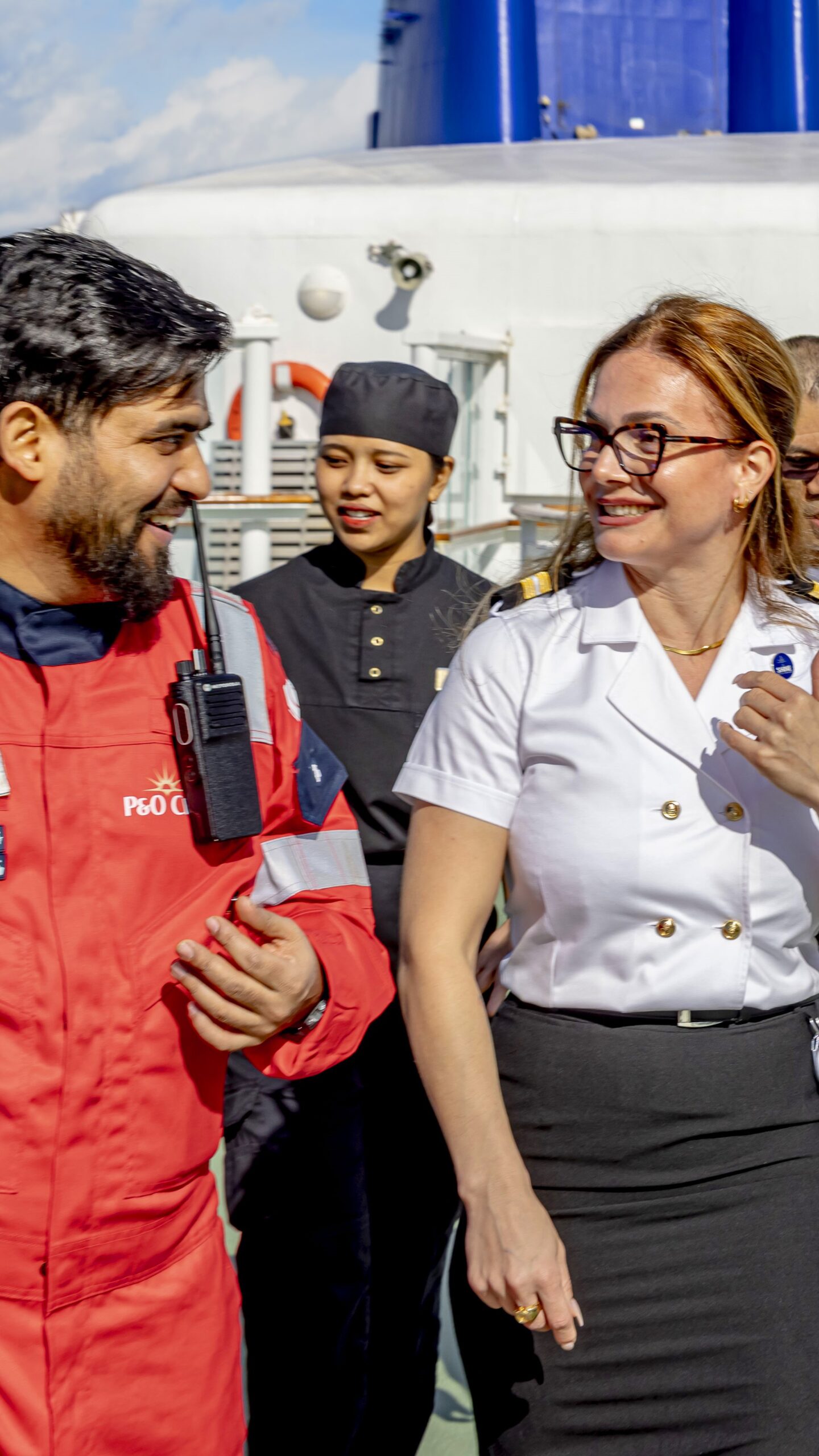 A man in red cruise ship overalls and a woman in a white shirt smile and talk whilst walking outdoors on deck, with another woman in a black uniform in the background