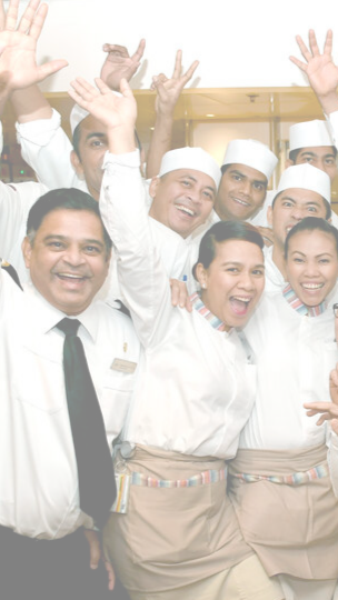 A group of smiling restaurant staff in uniforms pose together, raising their hands and appearing cheerful in a brightly lit setting, showcasing the teamwork found in cruise ship restaurant jobs.