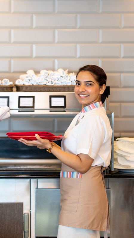A smiling waitress in uniform holds red plates in a restaurant setting, evoking the welcoming atmosphere often found in cruise ship restaurant jobs, with stacked serviettes and labelled containers visible near the counter in the background.