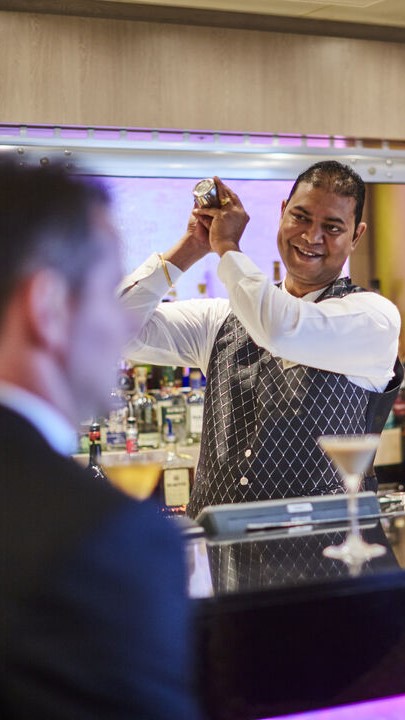 A smiling barman in a waistcoat and white shirt shakes a cocktail shaker behind the bar, showcasing the lively atmosphere typical of cruise ship bar jobs. Bottles and a martini glass are visible as he prepares a drink for a customer in the foreground.