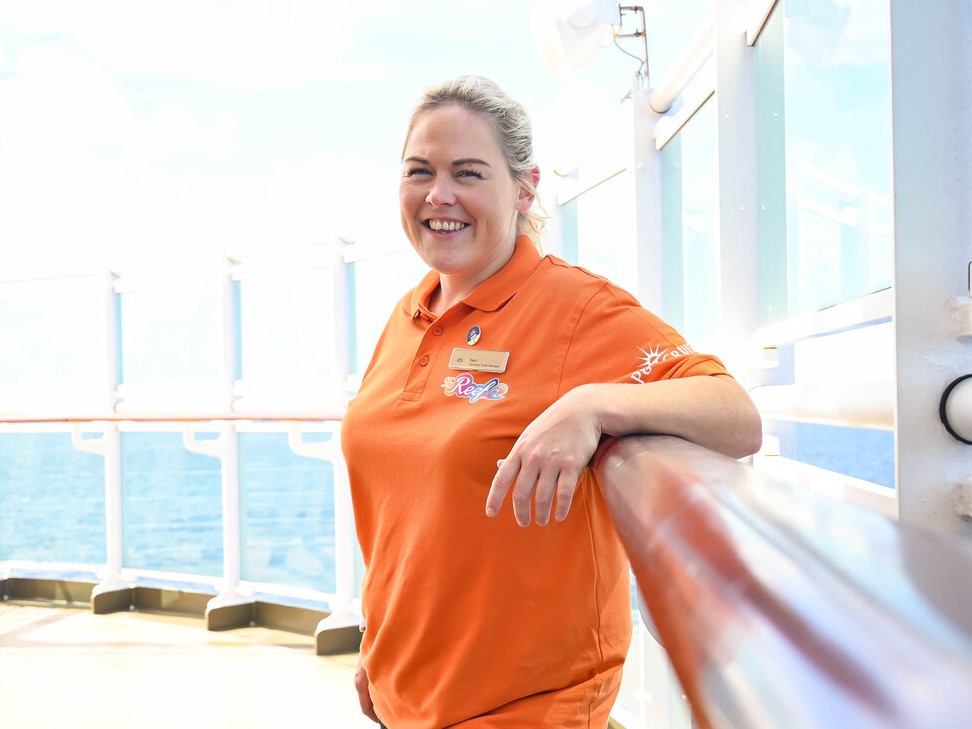 A woman with blonde hair in a ponytail, wearing an orange polo shirt with a name badge, stands and smiles on the deck of a ship with the sea and a blue sky in the background.