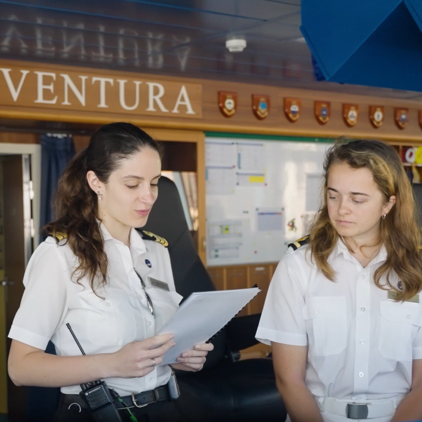 Two female officers in crisp white uniforms stand indoors; one reads from a clipboard whilst the other listens. Nautical decor and a “VENTURA” sign set the scene, highlighting P&O Cruises deck careers in action.