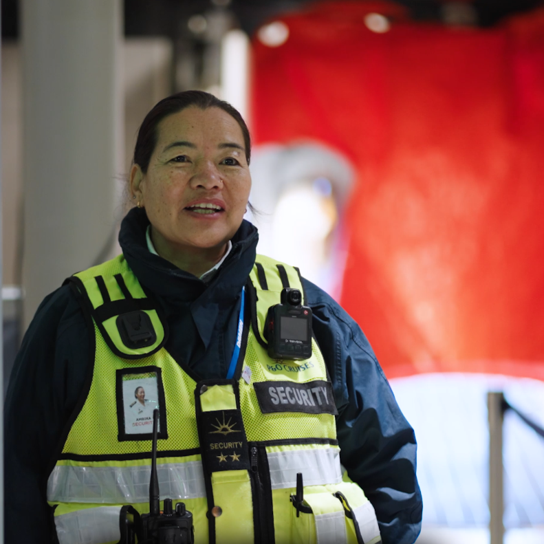 A security officer wearing a reflective yellow vest and radio equipment stands indoors, with a red background behind her. She has long dark hair pulled back and is looking slightly to the side, smiling—an example of P&O Cruises deck careers in action.