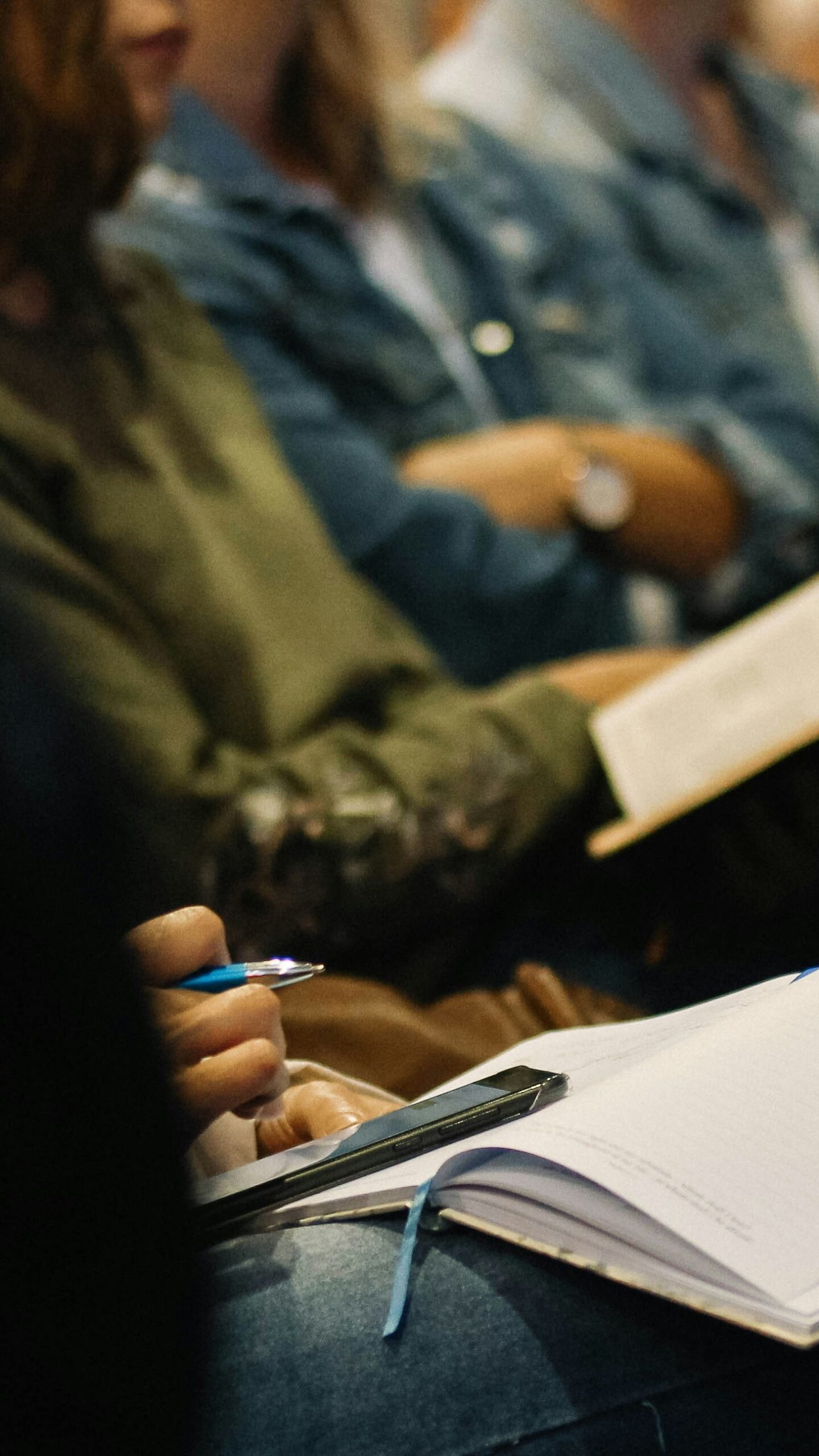 Close-up of people sitting with notebooks and pens, taking notes during what appears to be a lecture or meeting. The focus is on hands writing and holding pens, with blurred people in the background.