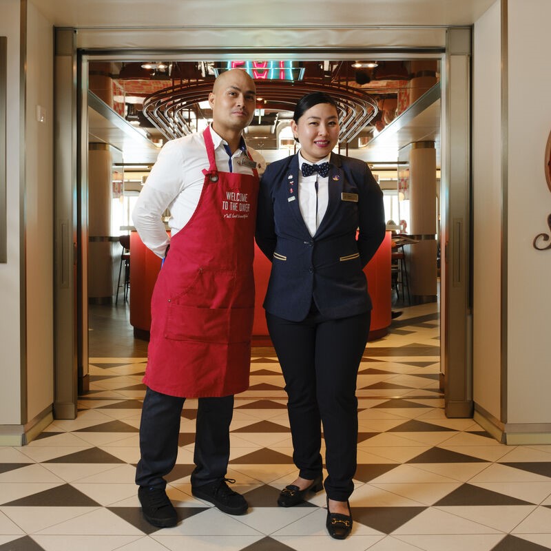 Two restaurant staff pose in a modern, well-lit dining area—showcasing the professionalism found in cruise ship restaurant jobs. One wears a red apron and white shirt; the other, a dark blazer and bow tie, stand on patterned tiles near the entrance.