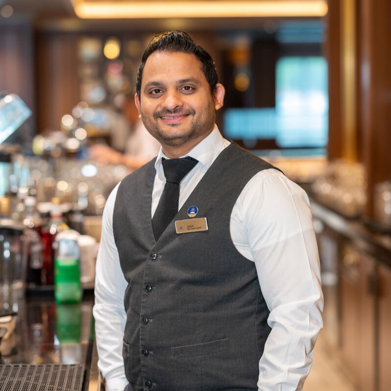 A smiling barman wearing a white shirt, black tie, and grey waistcoat stands behind a bar with bottles and glasses—capturing the elegance of cruise ship bar jobs in a warmly lit, sophisticated setting.