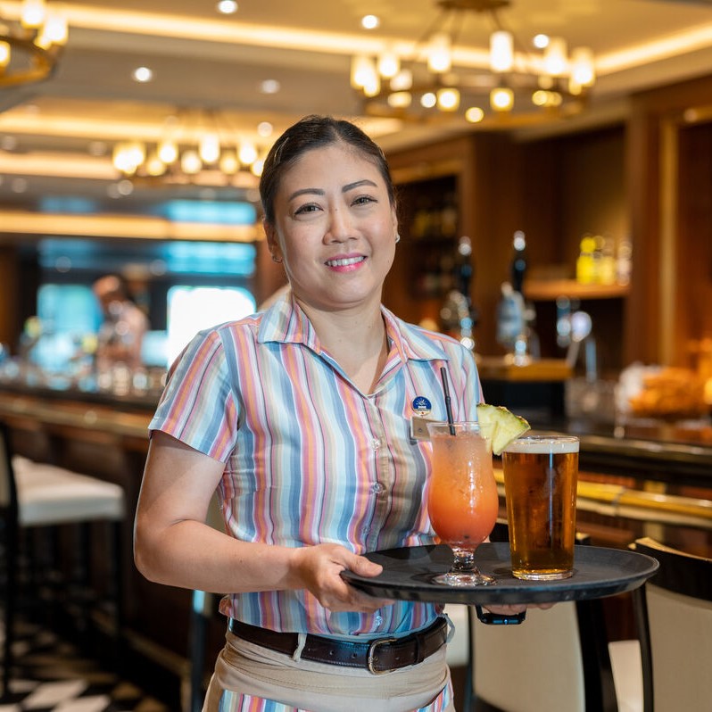 A smiling waiter in a striped shirt holds a tray with a cocktail garnished with pineapple and a glass of beer, standing in a stylish cruise ship bar with warm lighting and a long counter in the background—perfect for those seeking cruise ship bar jobs.