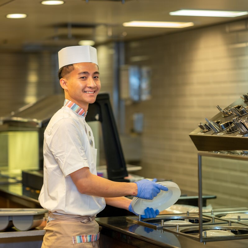 A smiling kitchen worker in uniform and gloves stands at a counter, holding clean plates in a commercial kitchen—an example of the teamwork found in cruise ship restaurant jobs, with cutlery and crockery visible in the background.