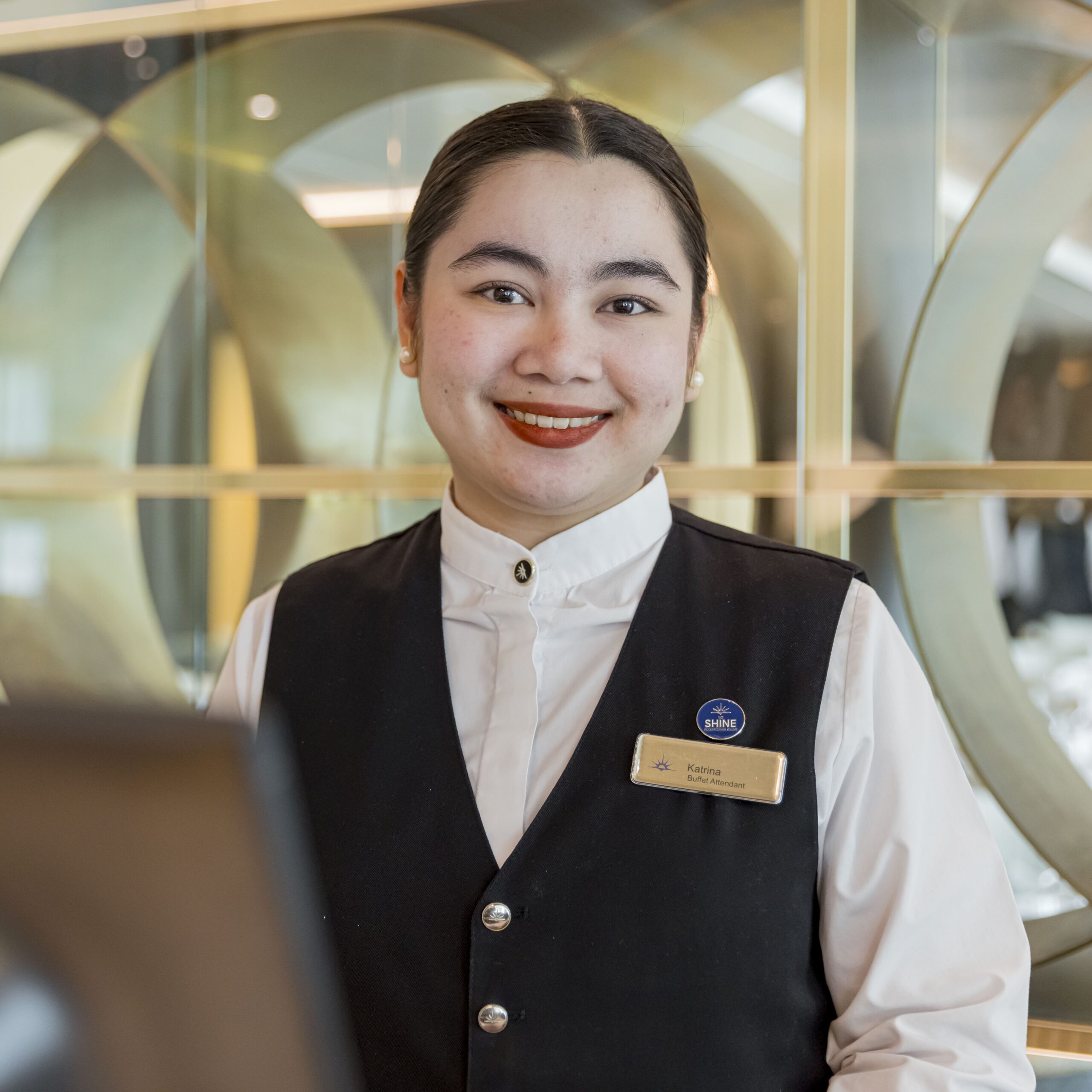 A smiling hotel staff member wearing a black waistcoat, white shirt, and name badge stands at a reception desk with a modern, elegant background—perfect inspiration for those seeking cruise ship restaurant jobs.