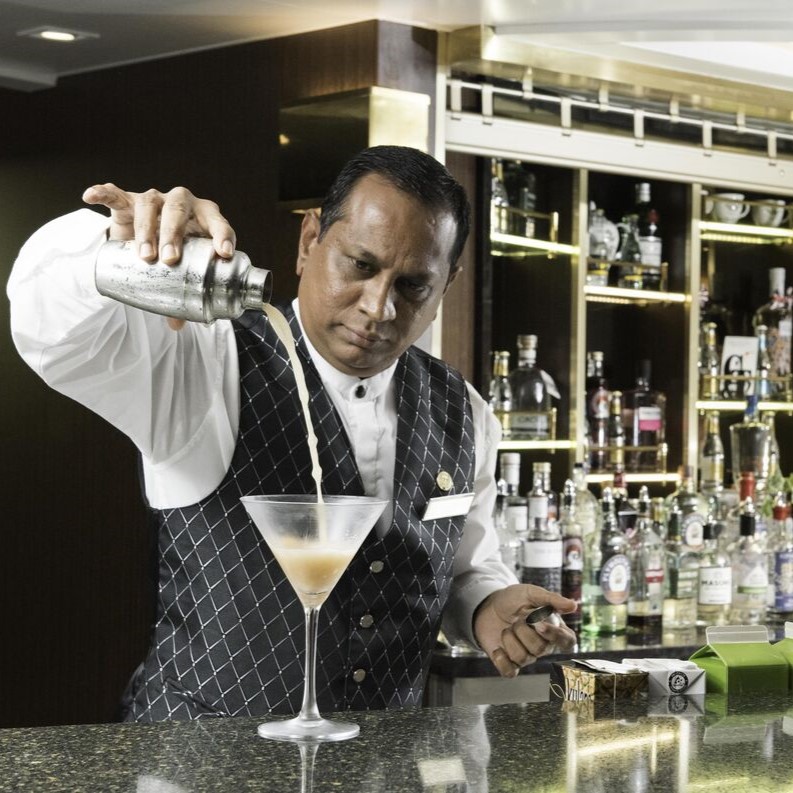 A barman in a black and white waistcoat pours a cocktail from a shaker into a martini glass at a bar, capturing the lively atmosphere often found in cruise ship bar jobs, with shelves of spirits bottles and bar tools in the background.