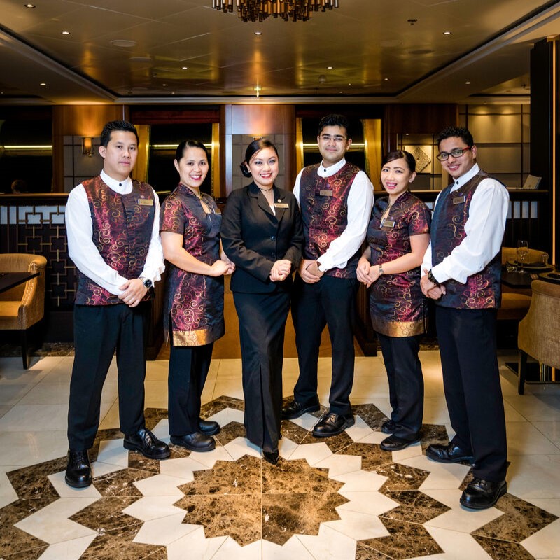 Six restaurant staff members stand together, five in matching patterned uniforms and one in a black suit, smiling and posing in an elegant cruise ship restaurant with marble floors and warm lighting. Perfect for those seeking cruise ship restaurant jobs.