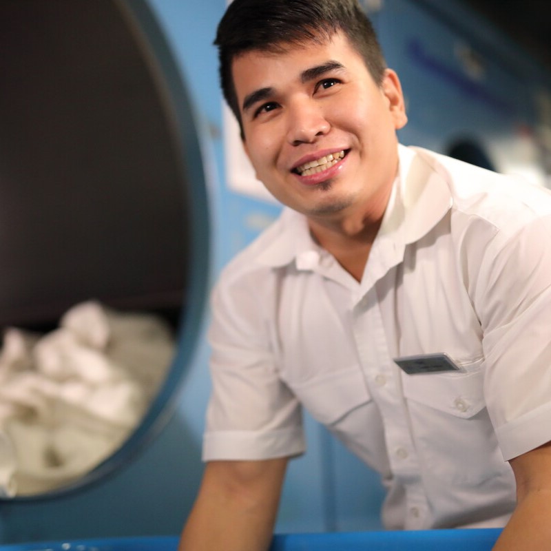 A man in a white uniform smiles whilst working in a laundry facility, standing in front of a large machine with a pile of white linen in the background.