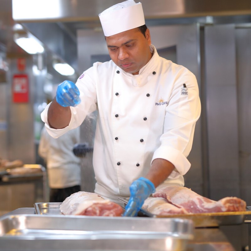 A chef in a white uniform and blue gloves sprinkles seasoning onto large pieces of raw meat on trays in a professional cruise ship galley kitchen.