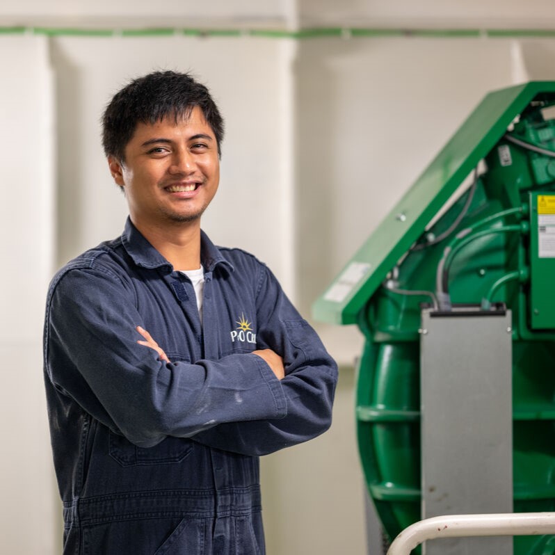 A man wearing a dark blue boiler suit stands with arms crossed, smiling, next to a large green industrial machine—reflecting the expertise and professionalism found in P&O Cruises technical careers—in a brightly lit indoor setting.