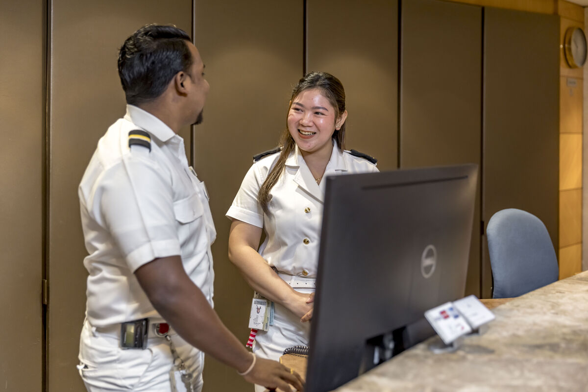 Two uniformed professionals, one man and one woman, stand smiling and conversing behind a reception desk with a computer monitor in front of them.