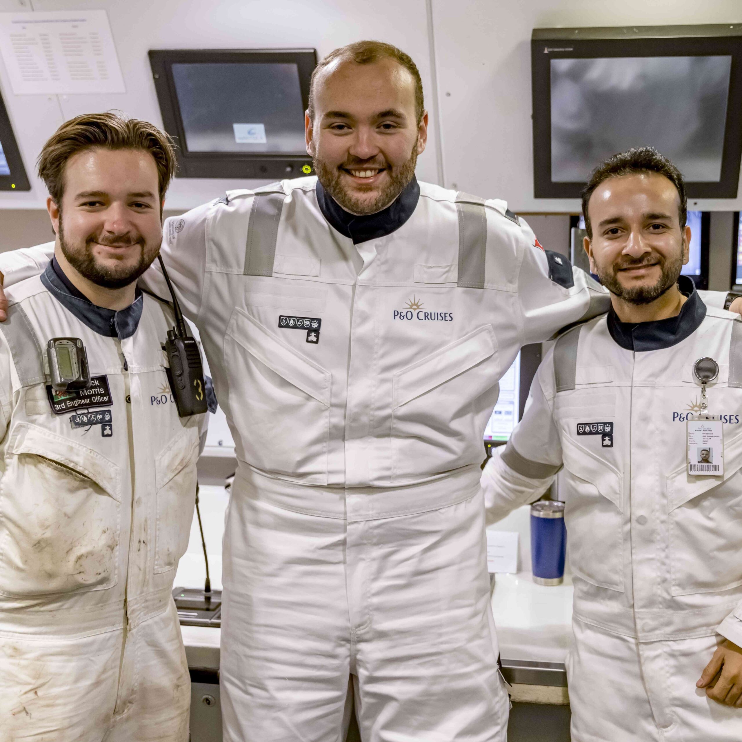 Three men in white P&O Cruises uniforms stand smiling together in a control room, surrounded by monitors and equipment—showcasing the teamwork and camaraderie found in P&O Cruises technical careers.