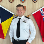 A smiling person in a white uniform shirt and black tie stands between two crossed maritime flags, with shield-shaped plaques displayed on the wall behind them.