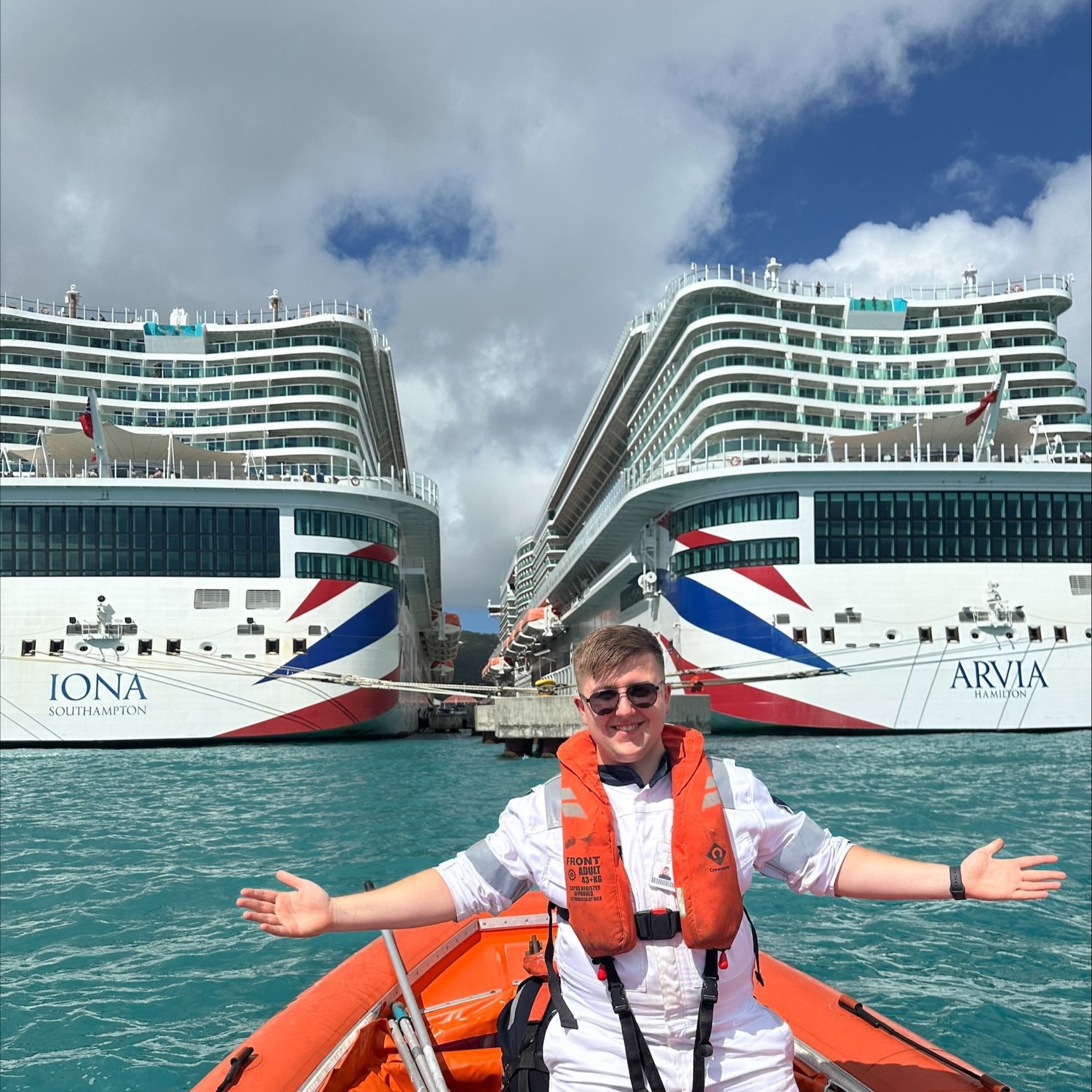 A smiling person in a lifejacket and white uniform, part of the Carnival Corporation cadet programme, sits in an orange boat with arms outstretched, posing before the Iona and Arvia cruise ships docked side by side in turquoise water.