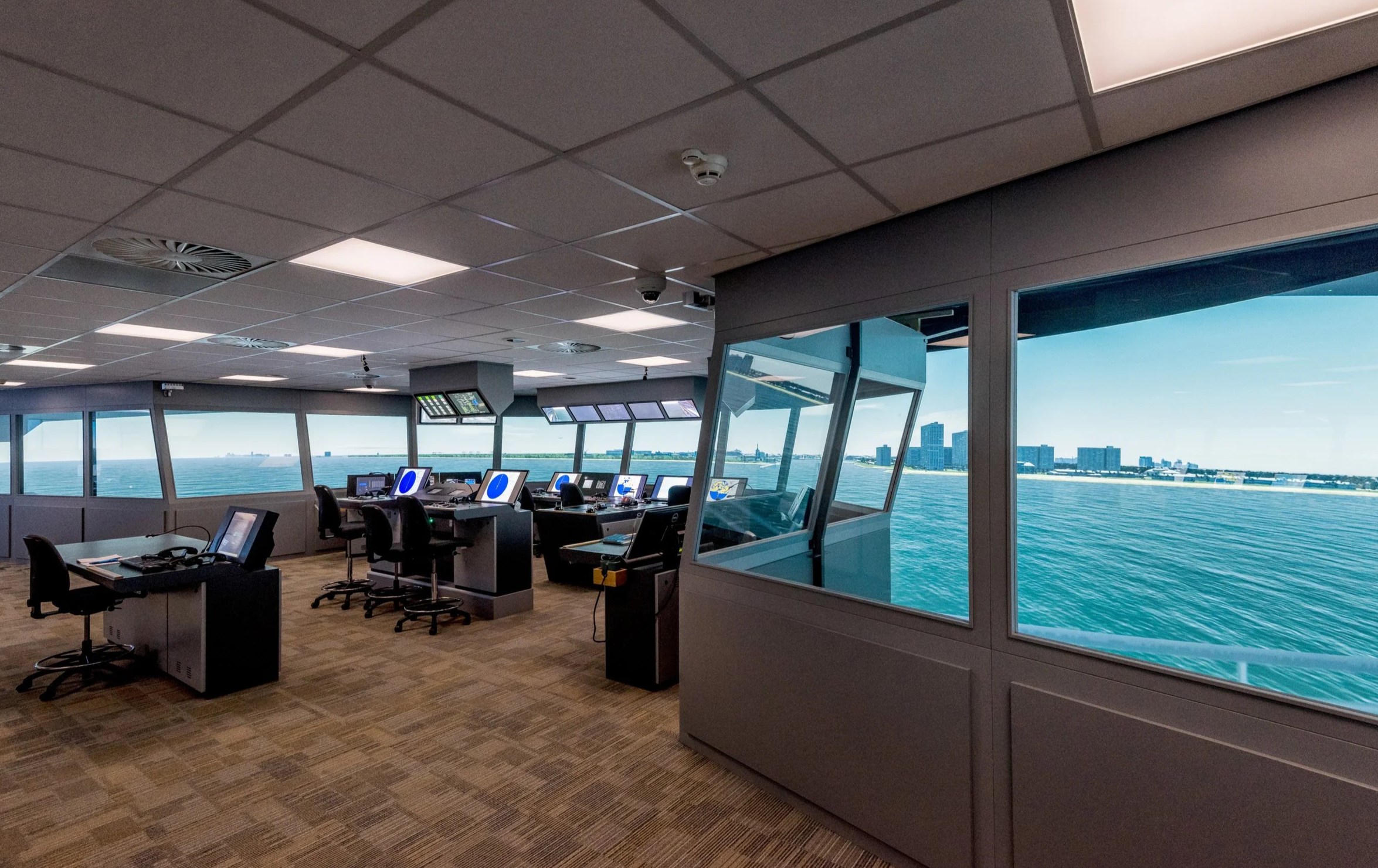 A modern ship bridge simulator room featuring multiple control stations, monitors, and large windows with a realistic sea view and distant city skyline—ideal for P&O Cruises technical careers training in a bright space with a tiled ceiling.