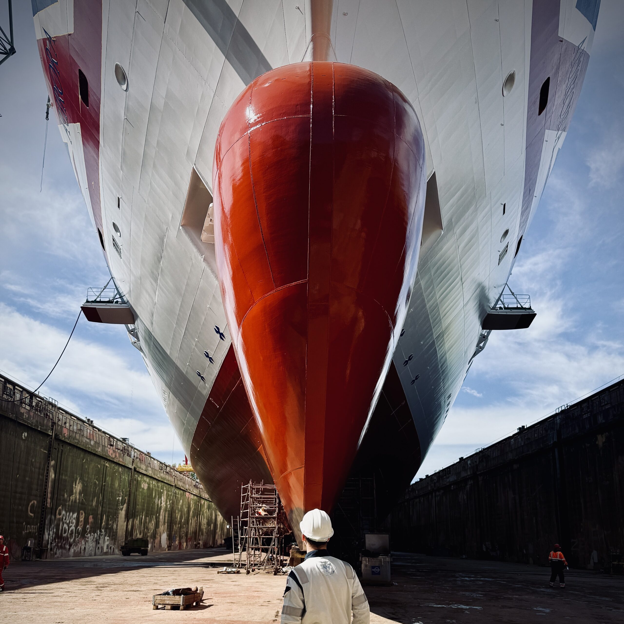 A person in a hard hat, possibly part of the Carnival Corporation cadet programme, stands in front of a large ship with a red hull in dry dock, looking up at the vessel. Scaffolding and another worker are visible beneath the ship.