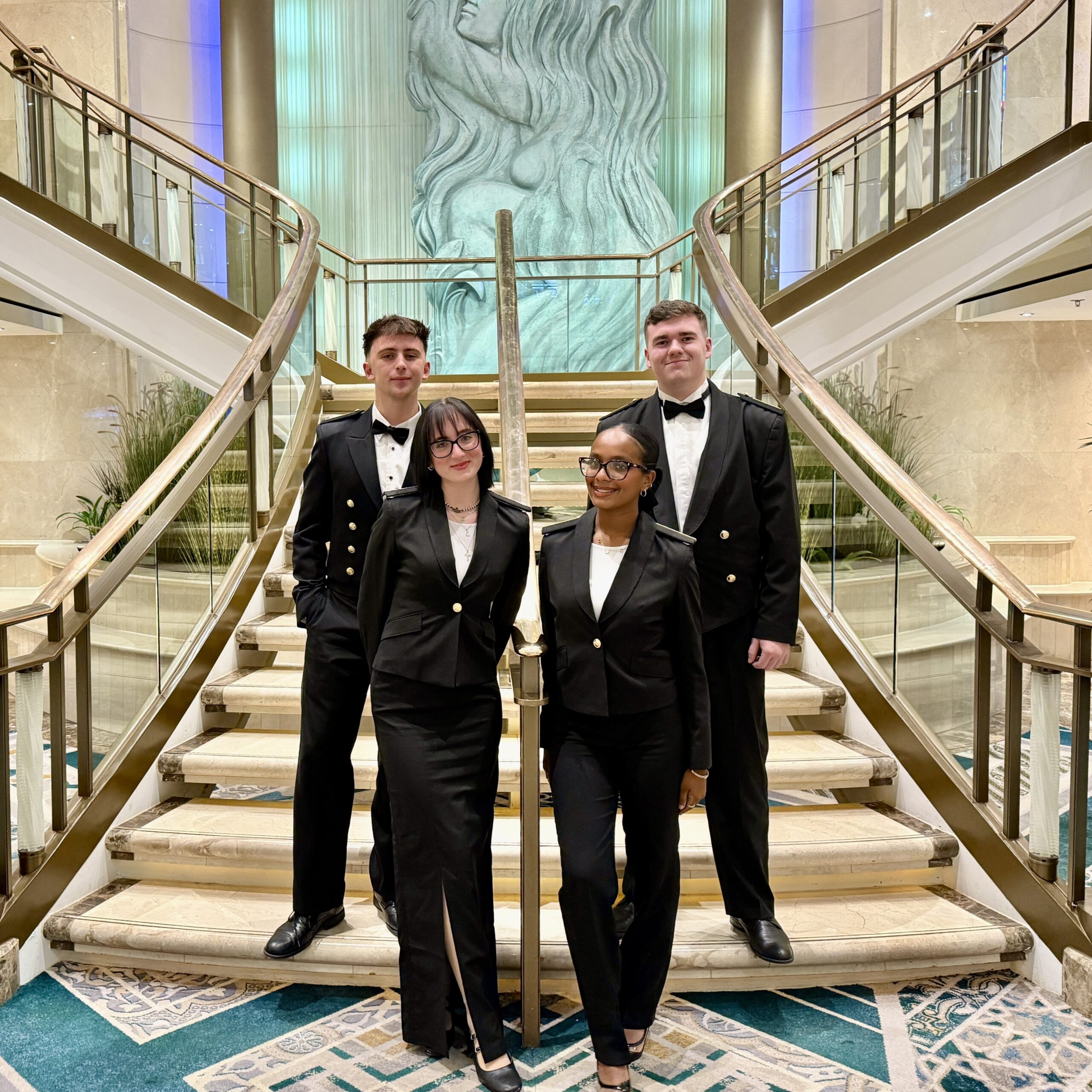 Four people in formal black suits and bow ties stand on an elegant staircase with glass balustrades in a luxurious lobby, highlighting the sophistication of the Carnival Corporation cadet programme, with a large sculpted wall art of a woman behind them.