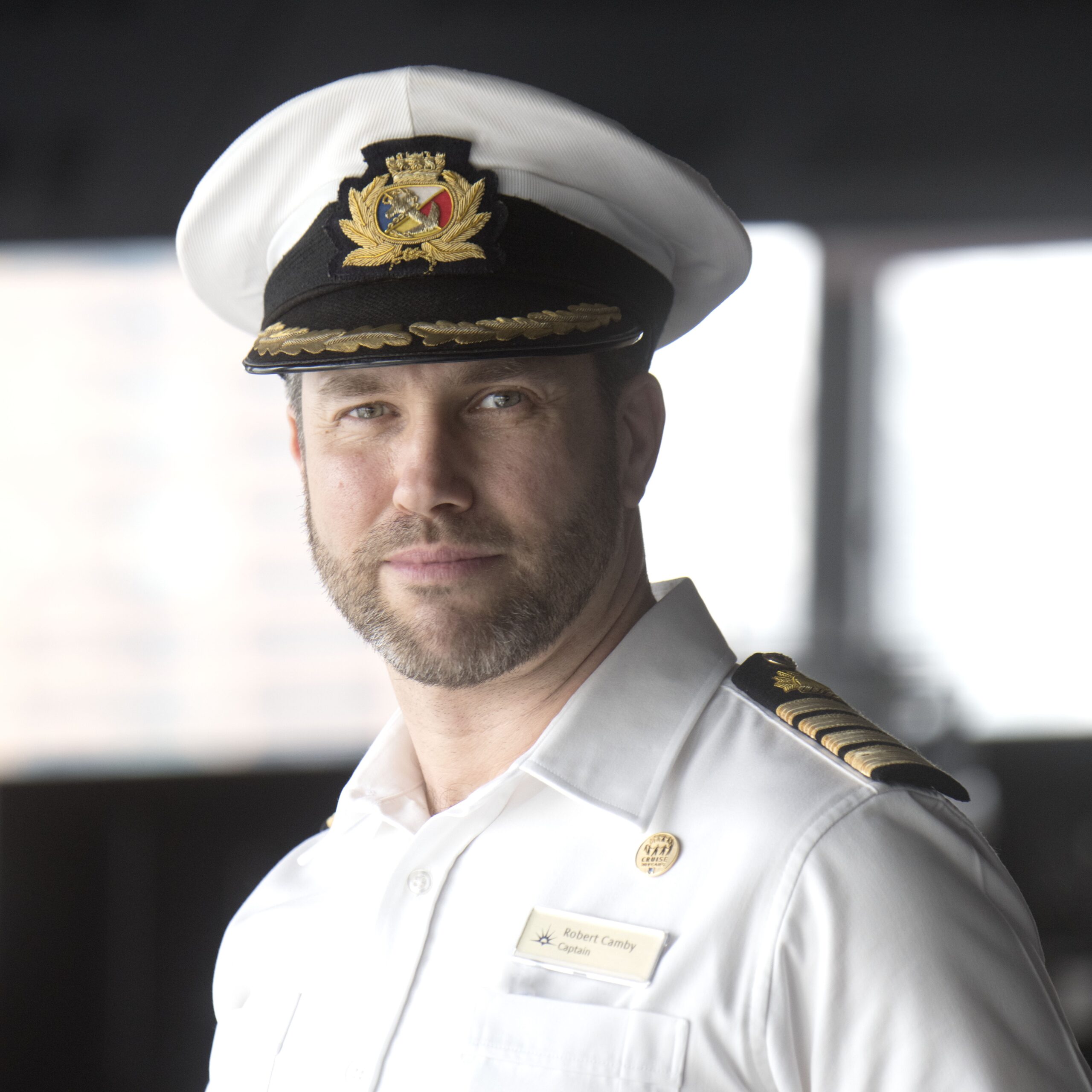 A man in a white naval captain’s uniform and hat, complete with name badge and epaulettes, stands indoors against a blurred background, representing the professionalism of P&O Cruises deck careers as he looks at the camera.