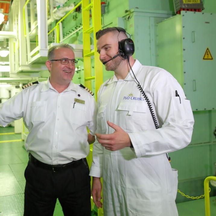 Two men in white uniforms, one with a headset, stand and talk in a brightly lit industrial setting with green walls and yellow railings—an environment typical for P&O Cruises technical careers. Both appear to be staff, possibly on a ship or in a technical facility.