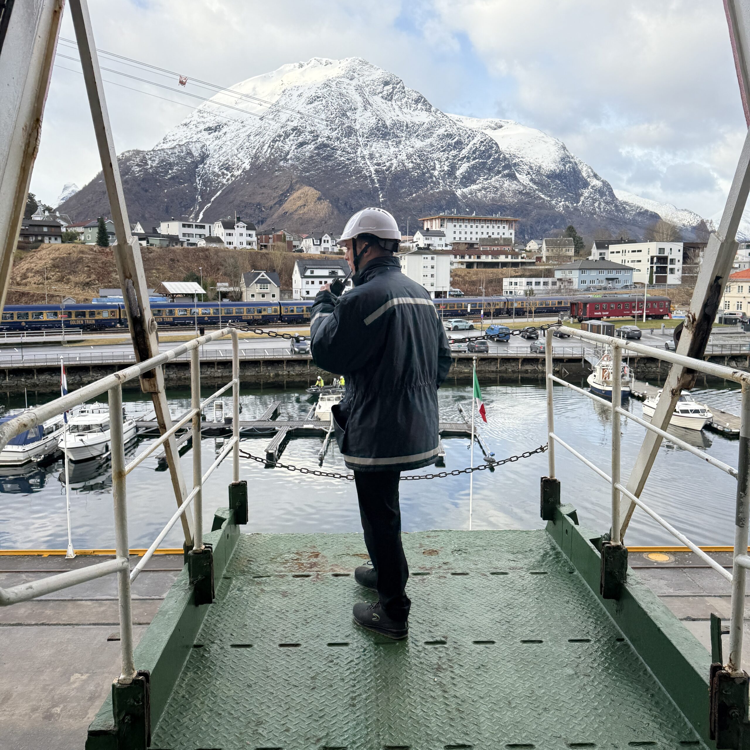 A person in safety gear, part of the Carnival Corporation cadet programme, stands on a metal platform overlooking a harbour with boats, buildings, and a snow-capped mountain in the background.
