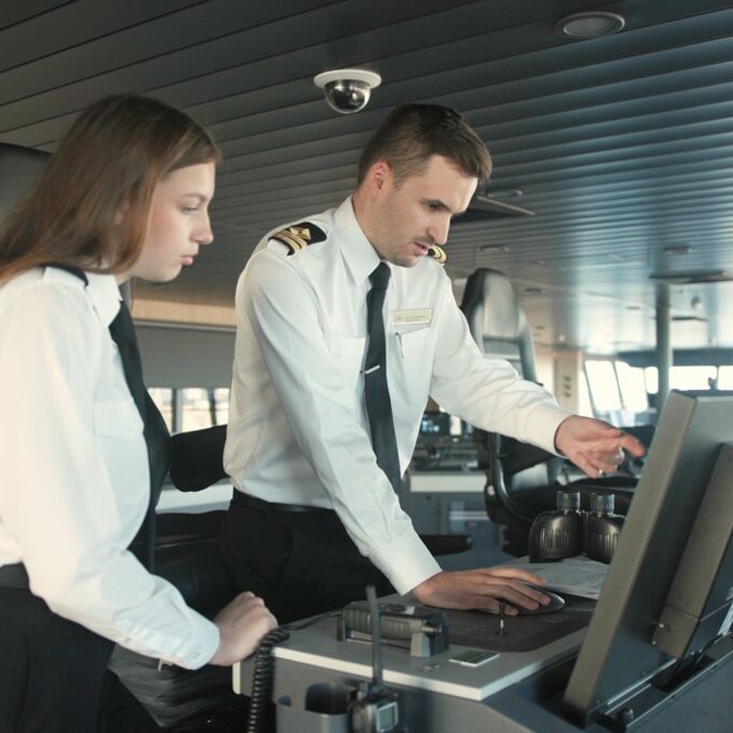 Two people in uniform stand at a ship's control panel. Part of the Carnival Corporation cadet programme, one points at the monitor whilst the other observes attentively, both focused on the screen in a modern ship's bridge.